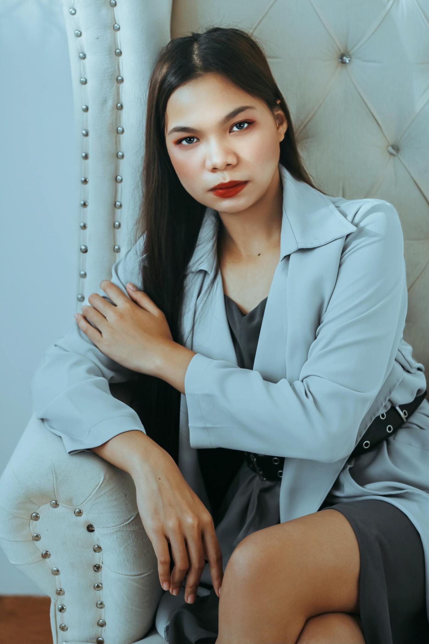 Portrait of a confident woman wearing a blazer, seated on a stylish chair, with a serious expression and red lipstick.