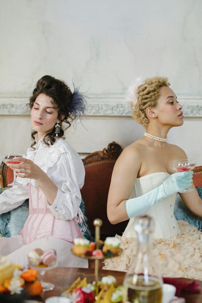 Two elegant women in period costumes enjoying drinks indoors, styled with vintage accessories.